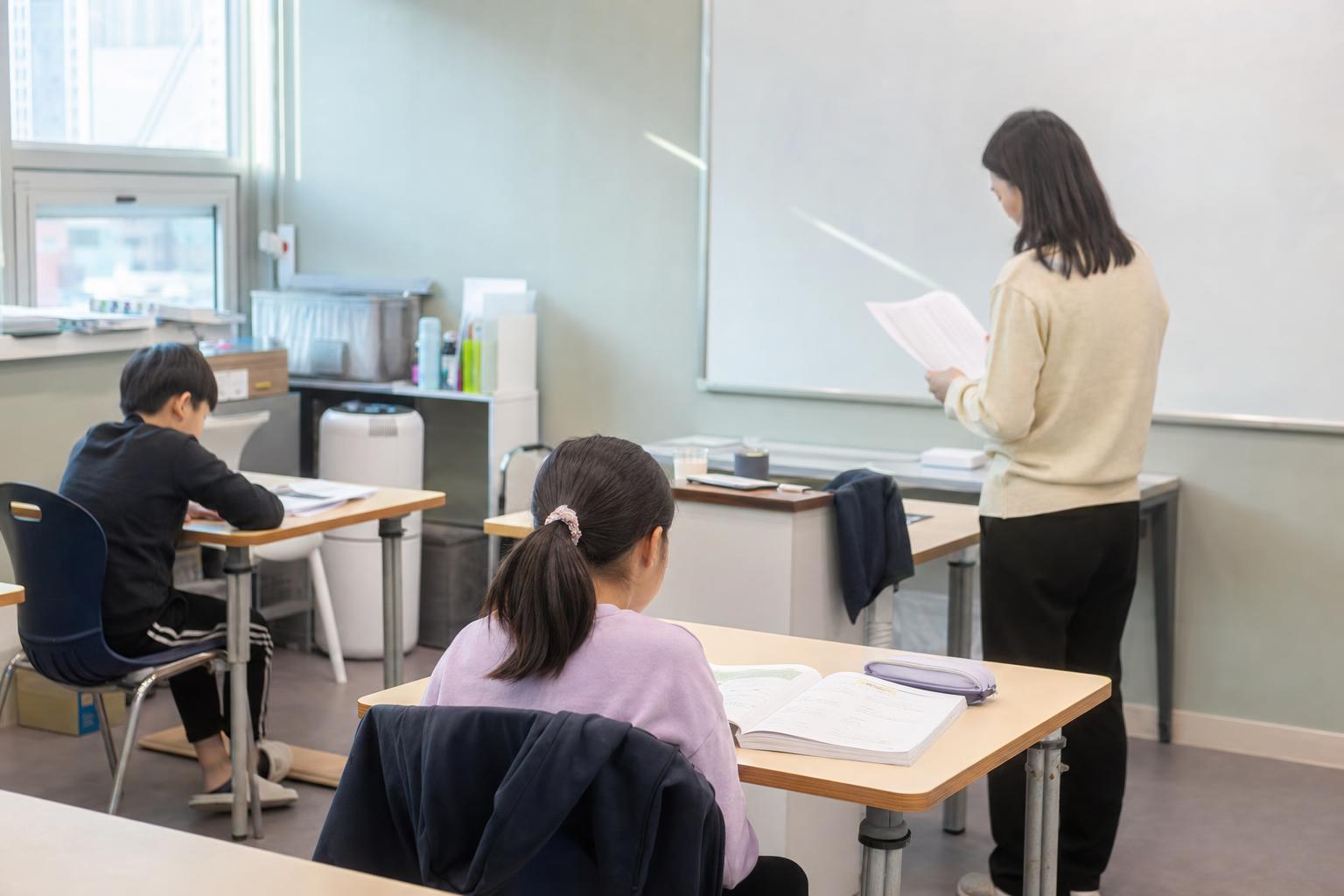 형제 학생이 학원 교실에서 각자 학습하고 강사가 출석과 수준 확인 자료를 보는 실사진형 장면